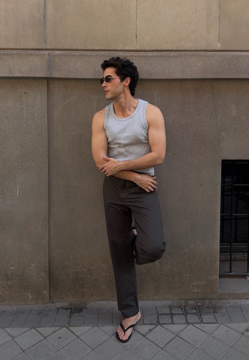 Homme portant des lunettes de soleil, un débardeur gris et un pantalon foncé, appuyé contre un mur beige avec un pied levé, debout sur un trottoir pavé.