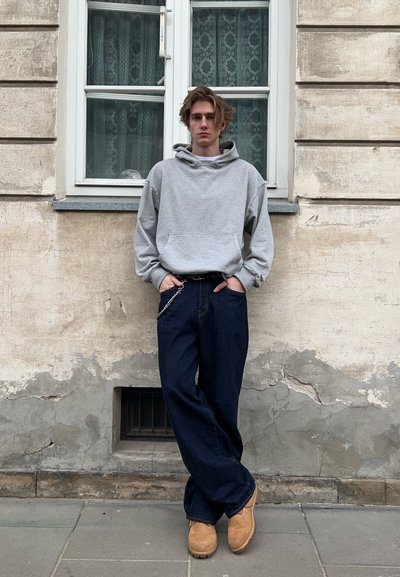 Sudadera gris con bolsillo tipo canguro, combinada con jeans sueltos de color azul oscuro y botas cortas en color marrón claro. De pie contra una pared texturizada.