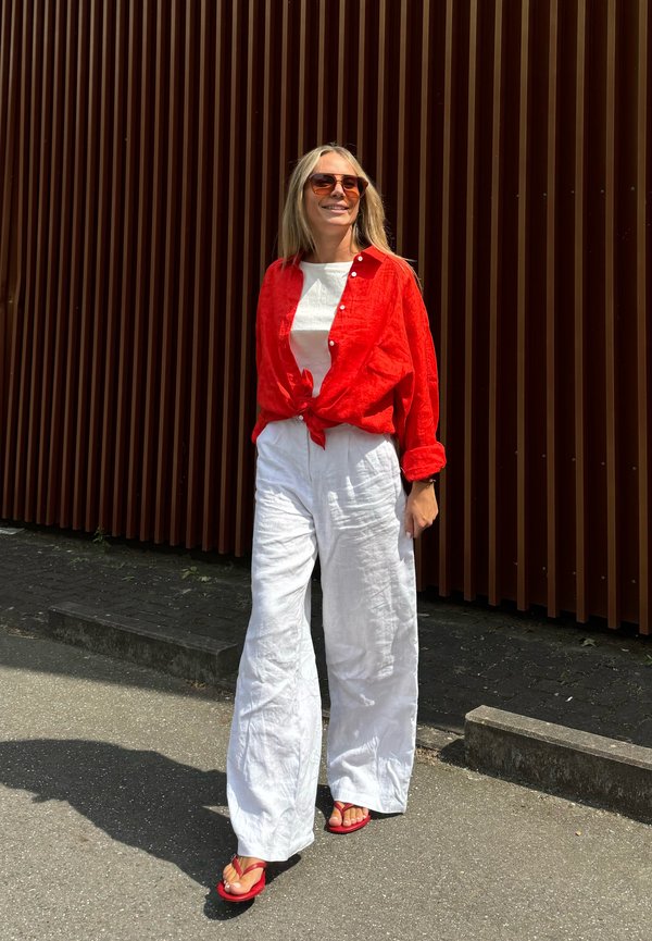 Frau geht draußen spazieren, trägt ein rotes Hemd, das in der Taille gebunden ist, weiße, weit geschnittene Hosen, rote Sandalen und Sonnenbrille vor einer braunen vertikalen Wand.