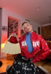 Blouson aviateur sportif rouge avec logo blanc, haut bleu, jupe noire avec motifs textuels, et sac à main plissé beige. Intérieur avec éclairage chaud.