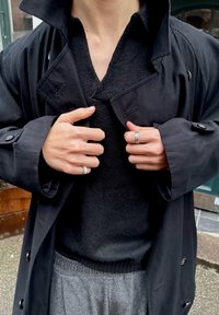 Trench-coat noir sur un pull en maille noir, pantalon de survêtement gris, les mains affichant plusieurs bagues en argent. Tissu texturé et détails des boutons visibles.