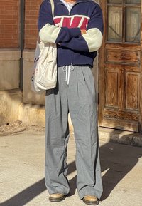 Σκούρο μπλε και κρέμα ζακέτα με φερμουάρ και κόκκινο γεωμετρικό μοτίβο, συνδυασμένη με ανοιχτό γκρίζο παντελόνι σε φαρδιά γραμμή και καφέ παπούτσια. Κρατάει μια μπεζ τσάντα ώμου.