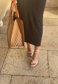 Καφέ τσαντάκι tote με γεωμετρικό σχέδιο και καφέ λεπτομέρειες, σε συνδυασμό με λευκές σανδάλες με λουριά, πάνω σε ένα άτομο που φορά μαύρο φόρεμα, το οποίο στέκεται σε πέτρινες πλακόστρωτες επιφάνειες.