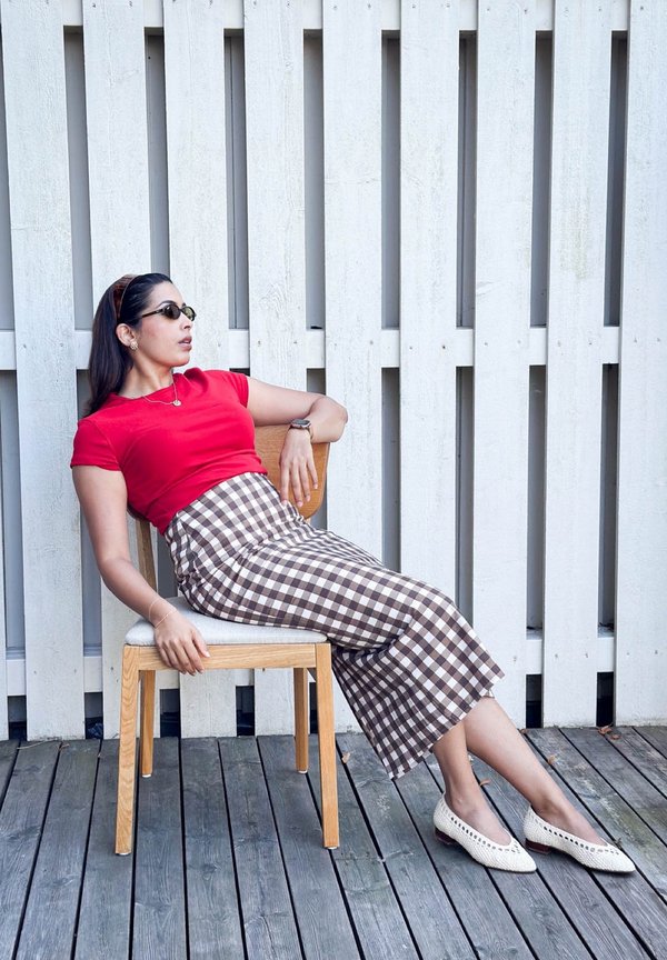 Femme portant un haut rouge et une jupe à carreaux, allongée de côté sur une chaise en bois contre un mur blanc à lames sur une terrasse en bois.