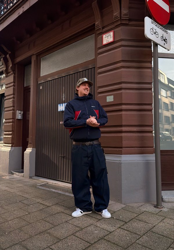 Vīrietis ikdienišķā apģērbā un kamuflāžas cepurē smaida, stāvot uz pilsētas ielas pie brūna stūra ēkas un ielu norādēm.