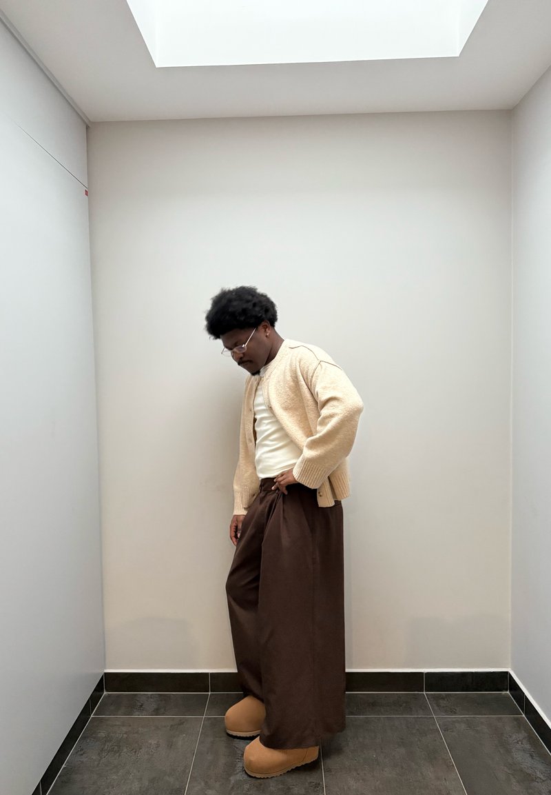 Cardigan beige chiaro sopra una camicia bianca, abbinato a pantaloni marroni larghi e zoccoli color cuoio, su uno sfondo di parete neutra e pavimento piastrellato.