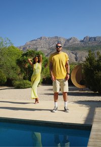 Duas pessoas estão perto de uma piscina. A mulher veste um vestido amarelo ajustado, enquanto o homem usa uma camisa amarela de mangas curtas e calções bege.