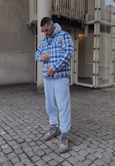 Chaqueta de franela a cuadros azul y blanca sobre una sudadera gris, combinada con pantalones de chándal azul claro y zapatillas grises, de pie sobre adoquines.
