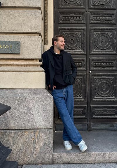 Blazer negro sobre una sudadera negra, jeans azules y zapatillas blancas. De pie junto a una pared de piedra texturizada con una puerta de madera detallada.