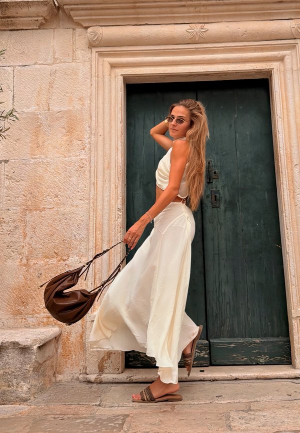 Vrouw met lang haar, gekleed in een witte jurk en sandalen, houdt een bruine tas vast en staat bij een stenen muur en een donkergroene deur.