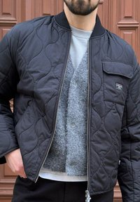 Czarna pikowana kurtka bomberka z zamkiem błyskawicznym, prążkowanym kołnierzem i przednią kieszenią; noszona na szarym swetrze dzianinowym. Materiał o fakturze, casualowy design.