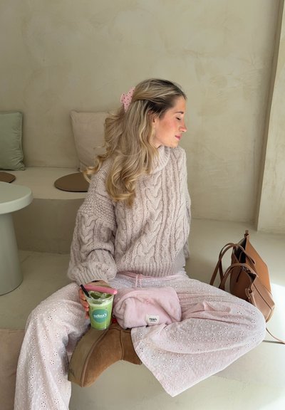 Jersey de punto en color rosa claro, pantalones estampados en rosa claro, botas de tobillo marrones, sosteniendo una bebida clara, con un bolso rosa y una bolsa marrón cerca.