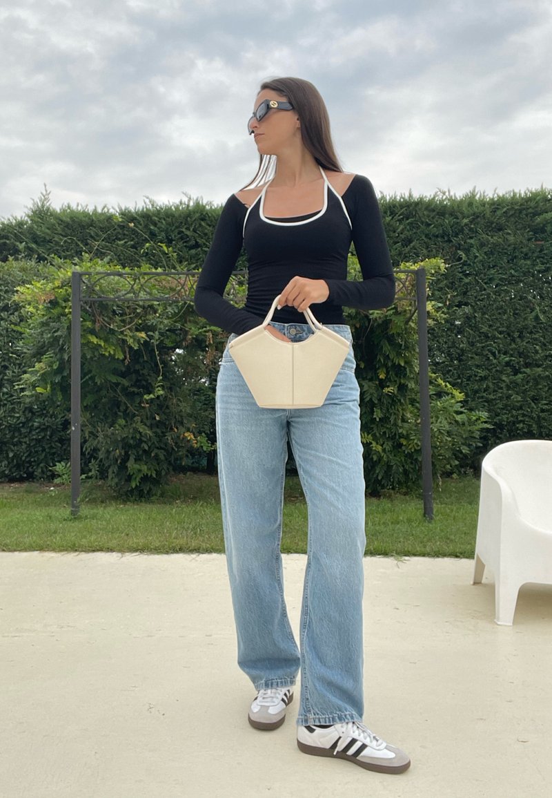 Borsa geometrica bianca in mano a una persona con una maglietta nera a maniche lunghe e jeans larghi blu chiaro, che indossa occhiali da sole scuri. Sfondo verde.