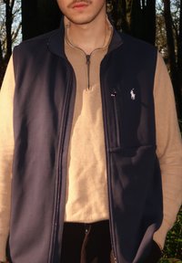 Veste sans manches bleu marine avec col montant, fermeture éclair et poche avant, portée sur un pull beige à finition texturée.