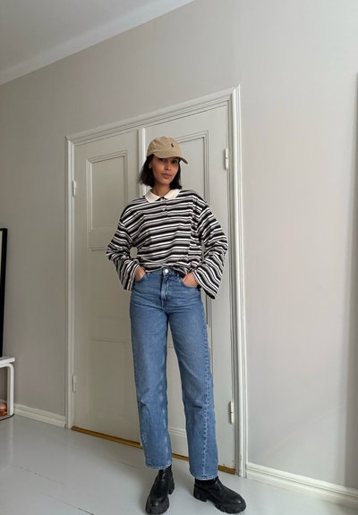 Camisa de rayas en blanco y negro de manga larga, con cuello de color claro, jeans azul de corte relajado y botas negras gruesas; se lleva una gorra beige.