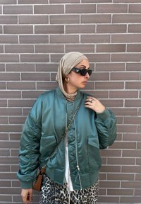Σατέν πράσινη bomber jacket με πλεκτούς καρπούς, δύο μπροστινές τσέπες, συνδυασμένη με μπεζ μαντίλι και παντελόνι λεοπάρ. Περιλαμβάνονται μαύρα γυαλιά ηλίου.