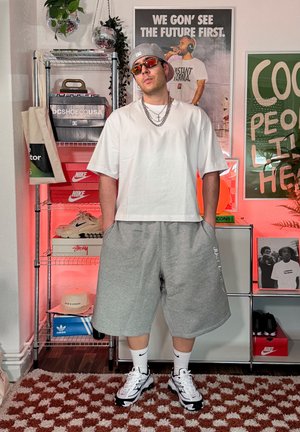 Hombre con gorra gris, gafas de sol rojas, camiseta blanca oversize, pantalones cortos grises, calcetines blancos y zapatillas en una habitación con pósters y estanterías para zapatos.