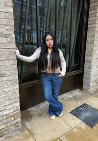 Weißer gerippter Cardigan über einem braunen Top, blau ausgestellte Jeans und beige Schuhe, posiert vor einer strukturierten Backsteinwand mit Glaselementen.