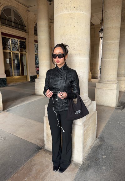 Mujer con chaqueta de cuero negra y gafas de sol sosteniendo auriculares blancos, parada junto a una gran columna de piedra en un arco.