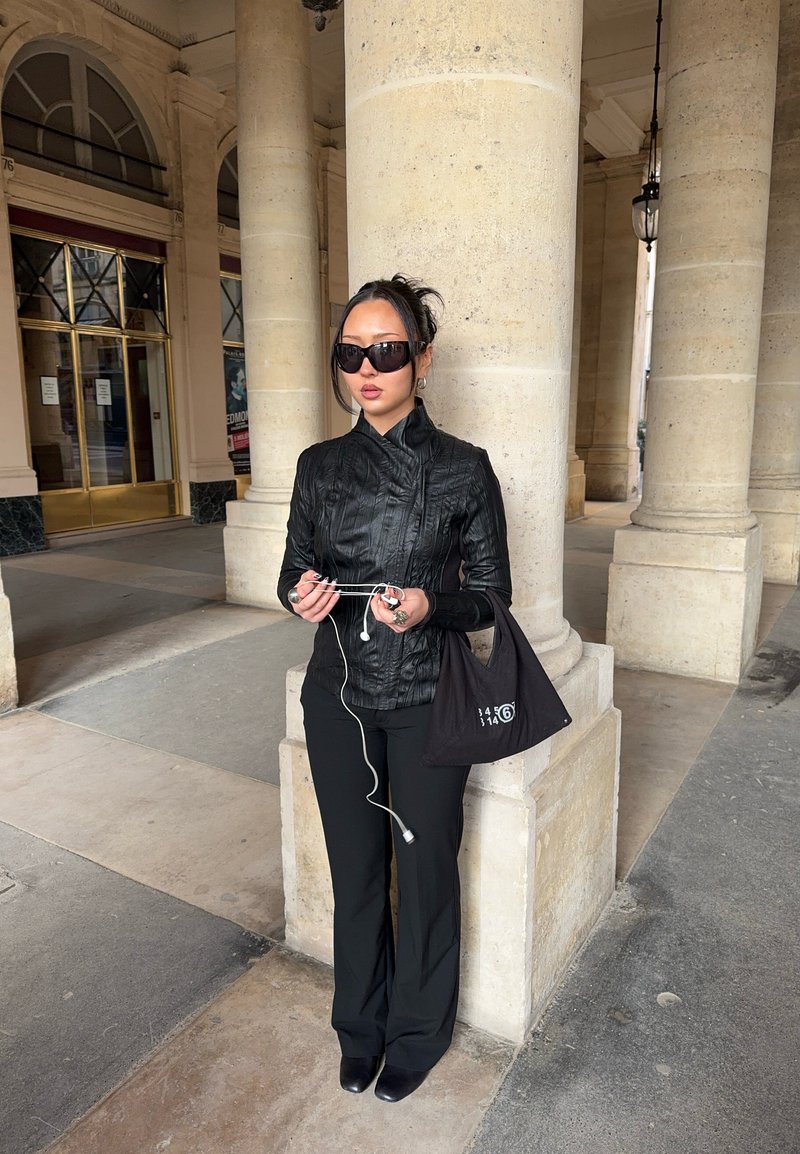 Femme en veste en cuir noire et lunettes de soleil tenant des écouteurs blancs, debout contre une grande colonne en pierre dans une arcade.