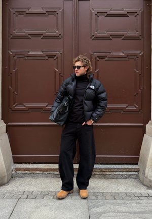 Hombre con chaqueta acolchada negra, pantalones negros, gafas de sol y botas marrones, de pie con una bolsa negra frente a una gran puerta marrón ornamentada.