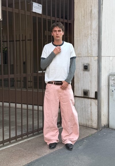 Joven parado junto a una verja de metal, con mangas largas en capas, pantalones rosas de gran tamaño y zapatos negros, tocándose el cuello y la cintura.