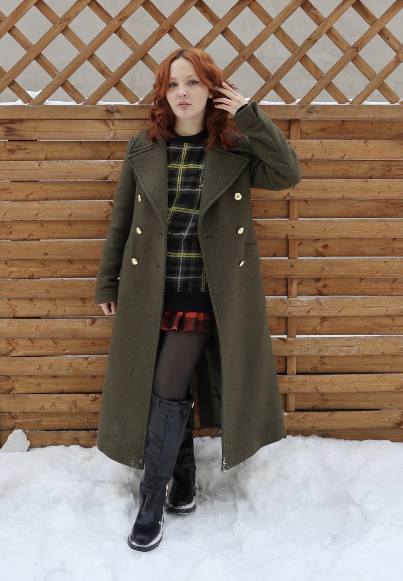 Femme aux cheveux roux debout dans la neige, portant un long manteau vert, une jupe à carreaux noirs et rouges, des collants noirs et des bottes noires montant jusqu'aux genoux, près d'une clôture en bois.