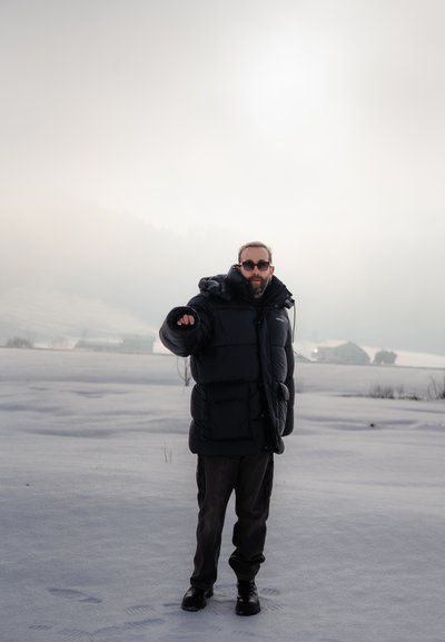Chaqueta de plumas negra con una gran capucha, textura acolchada y gruesa; llevada sobre pantalones oscuros y botas negras, de pie en un paisaje nevado.