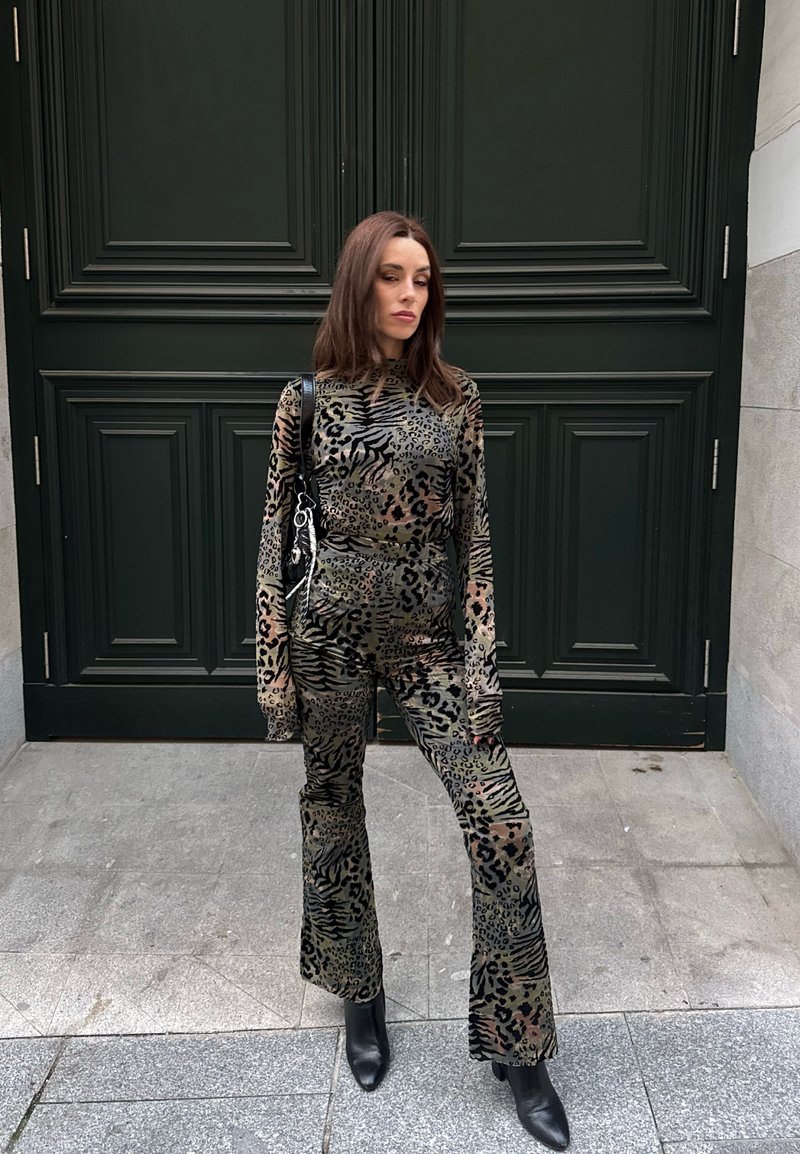 Donna con un top a maniche lunghe e pantaloni svasati coordinati con stampa animalier, stivaletti neri alla caviglia, in piedi davanti a una grande porta verde scuro.
