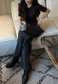 Camicia aderente nera, pantaloni grigi a righe larghe, stivaletti neri, tiene in mano una tazza di ceramica maculata, seduta su un divano in pelle bianca.