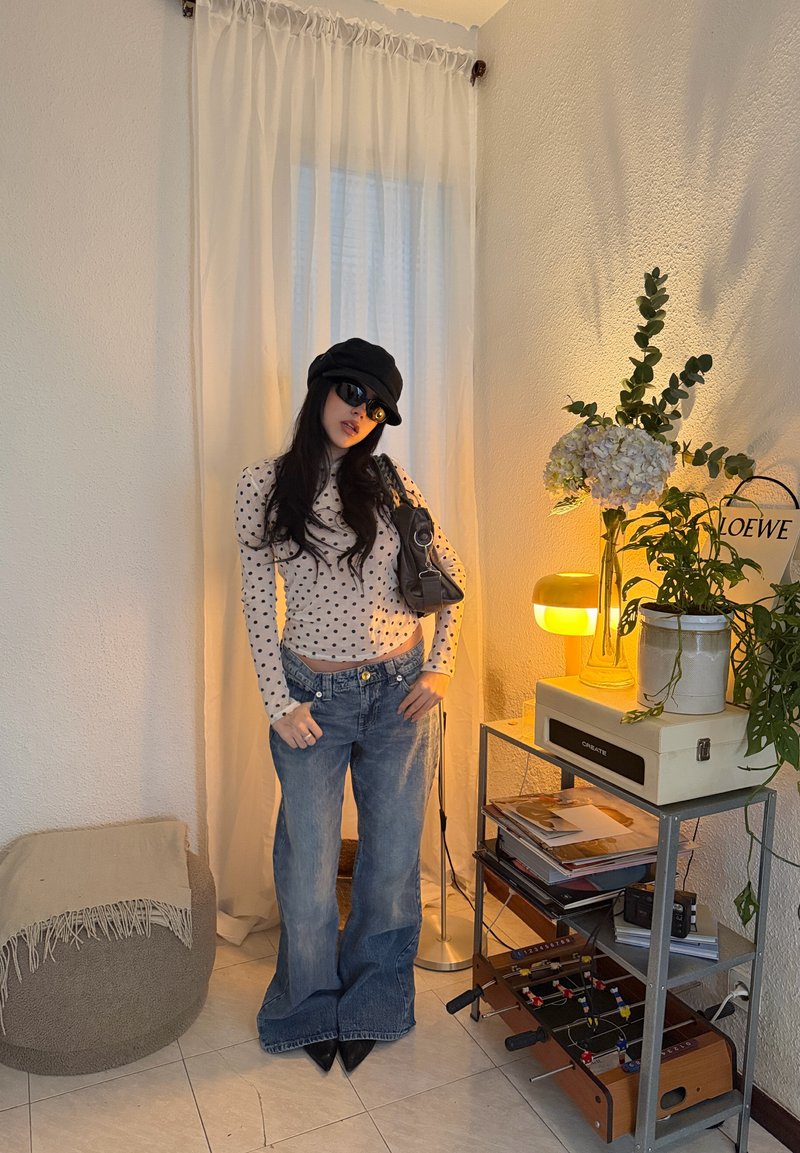Femme portant une casquette noire, des lunettes de soleil, un haut à pois et un jean, debout à l'intérieur près d'une petite table avec des plantes, une lampe et un baby-foot.