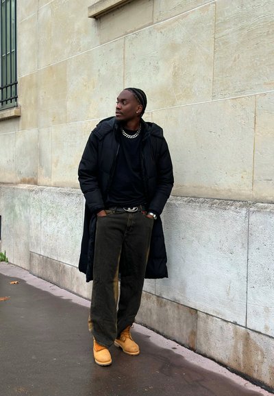 Abrigo acolchado negro, camiseta negra, pantalones verde oliva de corte holgado, botas de trabajo marrones con detalles en naranja, collar de cadena plateada, posando contra una pared.