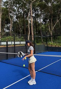 Joueuse de tennis en chemise grise et jupe plissée blanche, tenant une raquette noir et blanc, avec deux balles de tennis jaunes, sur un court bleu.