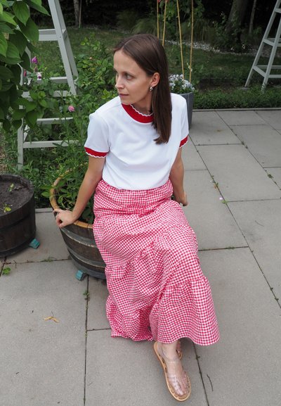 Camiseta blanca con escote y mangas rojas con ondas, combinada con una falda larga a cuadros rojos y blancos. Sandalias transparentes. Entorno de jardín.