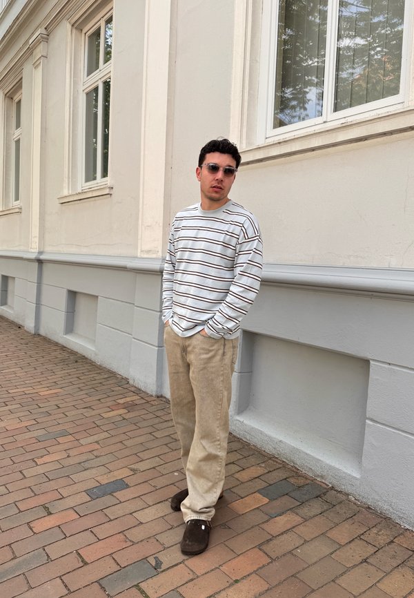Jeune homme portant des lunettes de soleil, une chemise rayée à manches longues, un pantalon beige et des chaussures foncées, debout sur un trottoir pavé près d’un bâtiment clair.