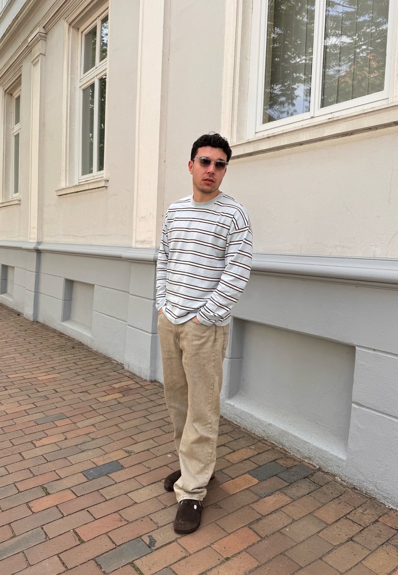 Jeune homme portant des lunettes de soleil, une chemise rayée à manches longues, un pantalon beige et des chaussures foncées, debout sur un trottoir pavé à côté d’un bâtiment clair.