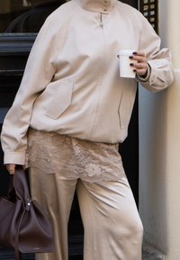 Giubbotto bomber beige con tasche laterali, indossato sopra un top in pizzo. Pantaloni beige a gamba larga, con in mano una tazza bianca e una borsa a tracolla marrone.