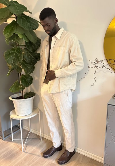 Camisa de algodón color beige y pantalones a juego con tejido texturizado. Zapatos marrones con un acabado suave. De pie junto a una gran planta verde.