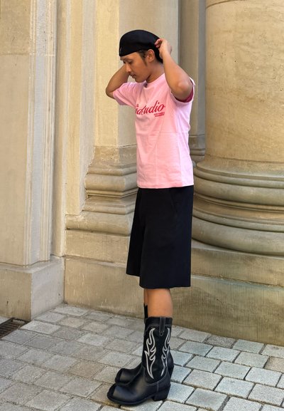 Camiseta de algodón rosa con texto rojo, pantalones cortos negros hasta la rodilla y botas vaqueras negras con patrones de llamas blancas, contra un fondo de piedra.