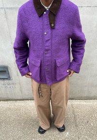 Personne portant une veste en laine violette avec un col marron, un pantalon large beige, des chaussures noires, et un porte-clés en perles accroché à la poche.