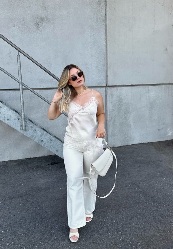 Femme en haut et pantalon blancs avec dentelle, portant des lunettes de soleil, tenant un sac à main blanc, debout sur l'asphalte près d'un escalier métallique et d'un mur en béton.