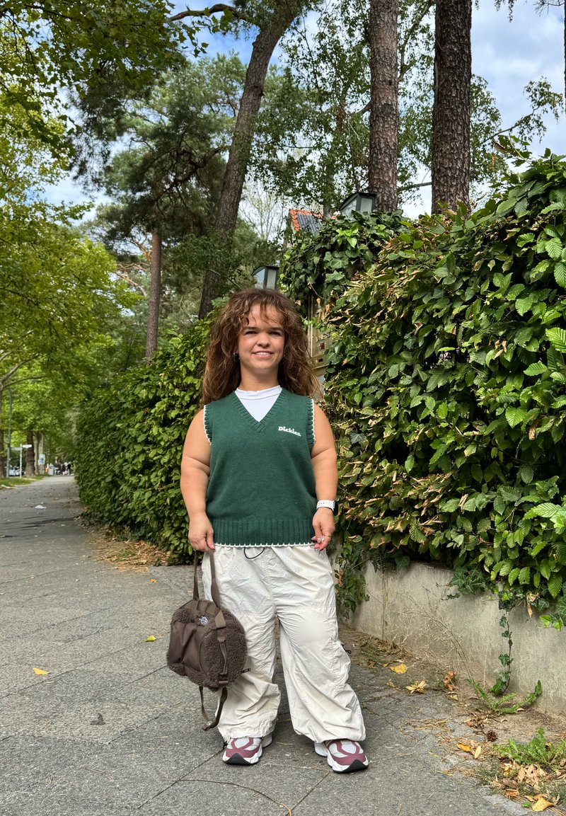Maglione verde a maglia con bordi bianchi a onda sopra una maglietta bianca, abbinato a pantaloni cargo crema larghi e uno zaino marrone in peluche, sneakers grigie.