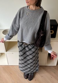 Persona con suéter gris acanalado, falda a cuadros en blanco y negro, zapatos marrones, collares dorados y llevando un bolso de hombro marrón oscuro en interiores.