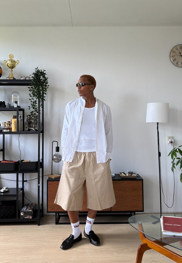 Person mit Sonnenbrille, weißem offenem Hemd, weißem Tanktop und beigen Shorts steht in einem modernen Wohnzimmer mit Regalen und einer Uhr an der Wand.