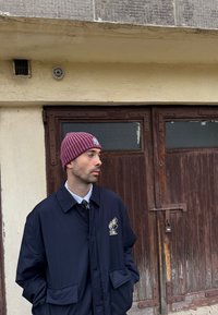 Tumši zila pogājama jaka ar logotipu, balta apkaklīte krekliņam un tumšsarkanā ribaina adīta cepure. Fonā brūnas koka durvis.