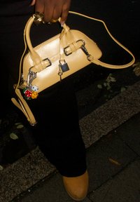 Sac à main en cuir jaune au design arrondi, avec des accents métalliques, un cadenas et des décorations de porte-clés colorés. Porté avec un pantalon noir et des chaussures marron.