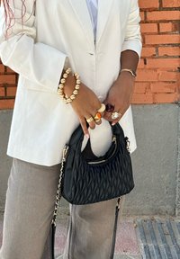 Black pleated handbag with a structured silhouette, gold hardware, and a detachable shoulder strap, being held by someone wearing a white blazer.