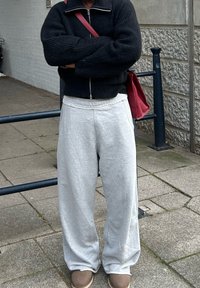 Maglione nero a zip, pantaloni sportivi grigi con tasche e calzature marroni. Una borsa rossa è appesa alla spalla. Ambientazione all'aperto.