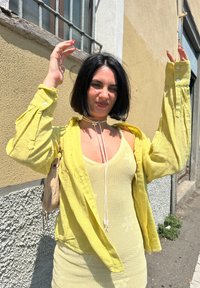 Żółta lniana koszula nałożona na pasującą dzianinową sukienkę. Szczegóły obejmują kwiatowy wzór na sukience oraz naszyjnik typu choker z akcentem w postaci kwiatu.