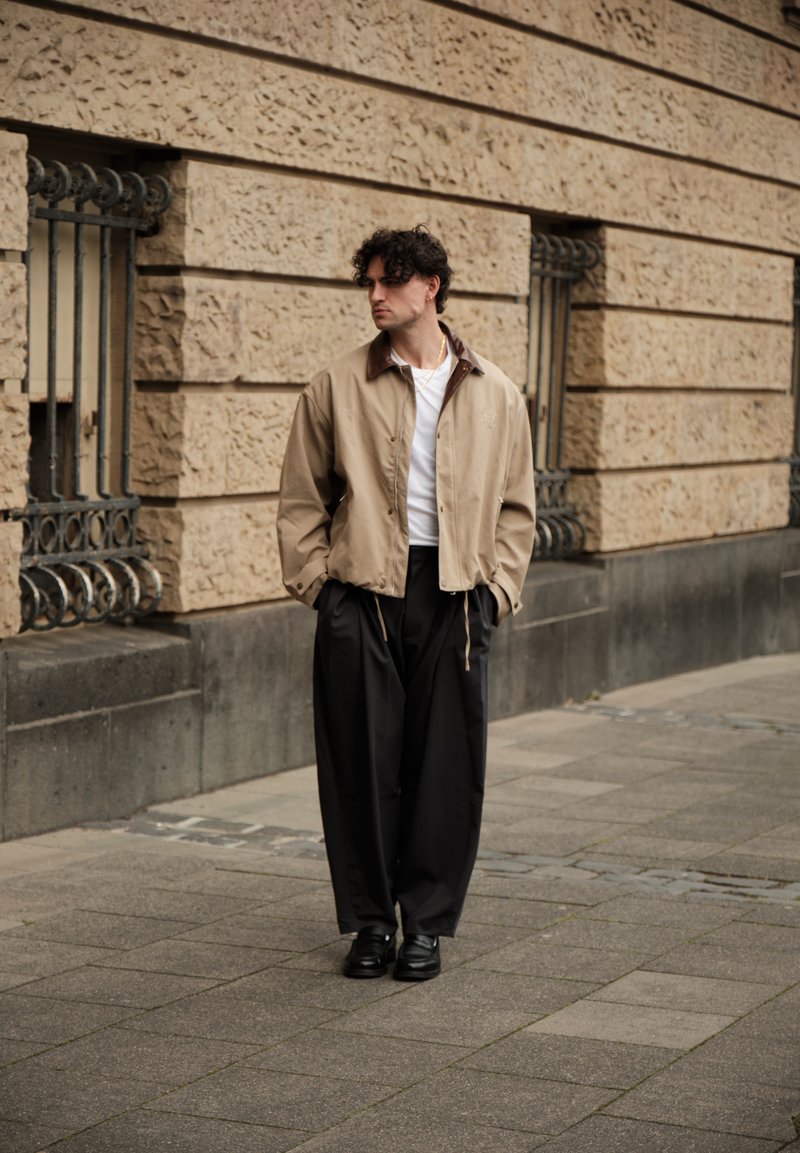 Άνδρας με κατσαρά μαλλιά στέκεται σε πεζοδρόμιο της πόλης φορώντας μπεζ μπουφάν, άσπρο πουκάμισο, φαρδιά μαύρα παντελόνια και μαύρα παπούτσια μπροστά από έναν υφασμάτινο τοίχο.